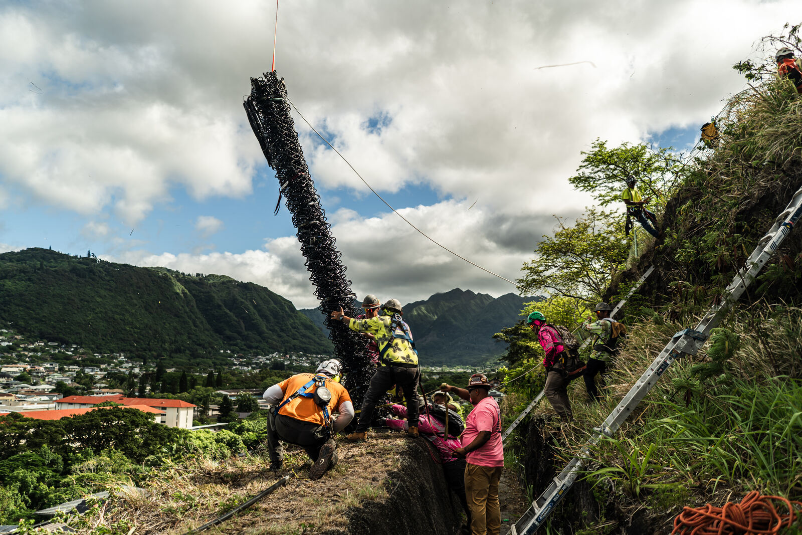 Crew guiding a rockfall barrier post under a helicopter above Manoa